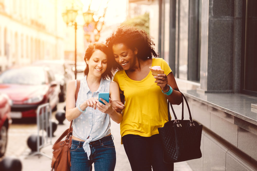 Two women walking down the road together. One is eating an ice cream while the other is pointing towards her phone screen